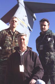 A man in a military uniform and two civilians stand near one another.  Behind them is the tail of a military aircraft.
