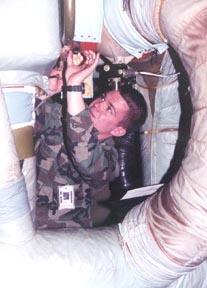A man in a military looks upward and inspects a box. he is surrounded by a large white flexible duct.