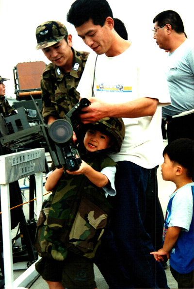 A small boy in a military uniform and helmet holds what appears to be a missile launcher.  His father, in civilian clothing stands behind him.  Nearby is an adult man in a military uniform. Another small boy watches.