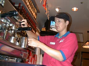 A woman in a pink shirt and a ball cap operates the milk steamer on an expensive-looking coffee machine.