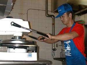 A man in a red shirt with a blue apron and ball cap operates a machine that makes a flat pizza crust.