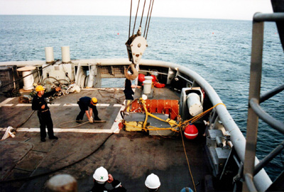 Two men and wreckage are visible on the deck of a ship.  The hook from a crane swings in from above.  The ocean is visible all around.