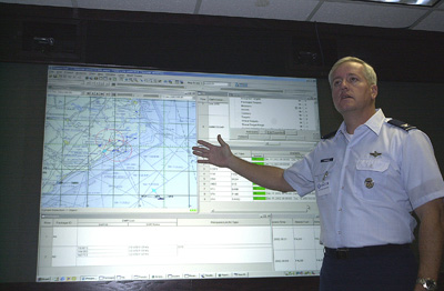 A man in a military uniform stands in front of a projection screen that displays a map and other information.  