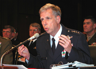 A man in a military uniform sits at a table and speaks into a microphone.  Other military personnel are seated behind him.