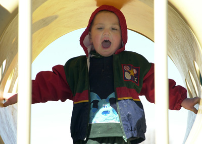 A small child in a red and blue coat stands inside a tube structure on a playground.