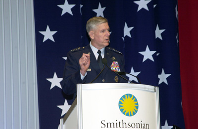 A man in a military uniform stands behind a lectern that has the word "Smithsonian" on it.  Behind him a large American flag hangs on the wall.