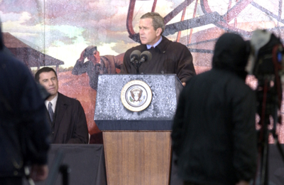 A man stands behind a lectern and speaks into a microphone. A mural is on the wall behind him, but it's not apparent what it depicts. Another man sits to the left.  Others stand and watch.