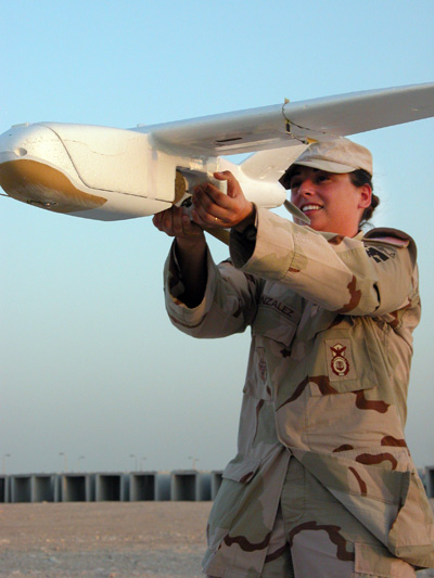 A woman in a military uniform holds a small foam aircraft in her outstretched arms.  She is outdoors in a desert environment. In the background are concrete structures.  