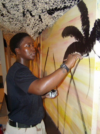 A young woman in a black T-shirt paints a black palm tree on a concrete wall.  Above her head is camouflage netting.
