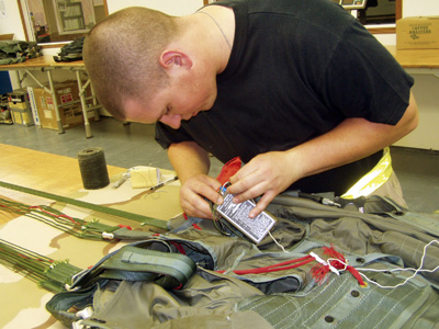 A man in a black T-shirt leans over a table. There is a parachute pack on the table. He holds an electronic device in his hands.