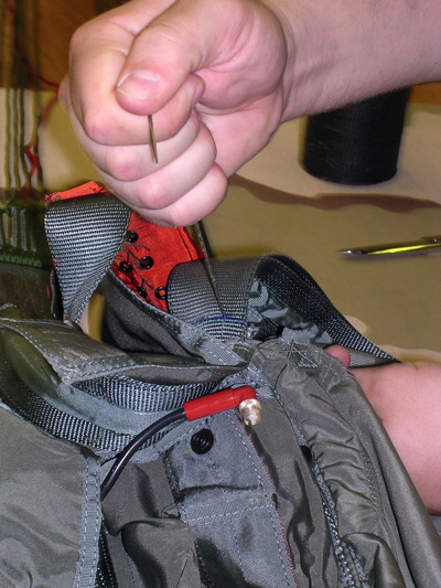 A hand holds a thick sewing needle and pulls thread through a strap on what appears to be a parachute pack.