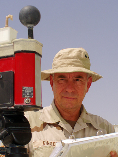 Outdoors, a man in a military uniform holds a clipboard and looks at a piece of weather-sensing equipment mounted on a tripod.