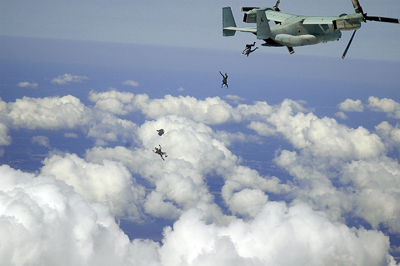 An aircraft flies high in the sky over clouds.  Four individuals jump out of the back of it.