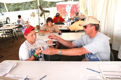 Under a tent, a boy in a red hat sits at a table.  He holds a model airplane. A man assists him.  Other boys sit at other tables.