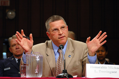 A man in a beige suit sits at a table and speaks into a microphone.  A pitcher of water sits next to him.  A sign says "Honorable Michael L. Dominquez." Others sit behind him.
