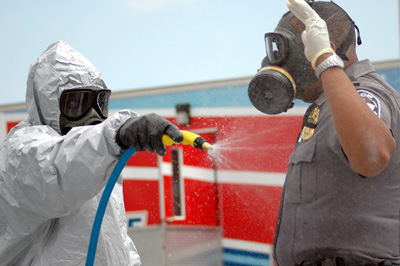 A person in protective gear uses a hose to spray a man in a police uniform.