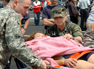In a parking lot, a woman in a military uniform tends to a woman on a stretcher who is covered in a blanket. A man in a military uniform assists.