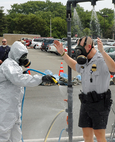 In a parking lot, a man in a police uniform is hosed down by a portable shower and a person in protective gear and a respirator. 