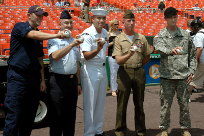 Five individuals in military uniforms stand together and each holds a baseball. In the background are hundreds of red seats. 