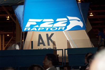 A cover, with the words "F22 Raptor," is lifted off the tail fin of a military aircraft to reveal the letters "AK."