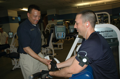 A man sits on exercise equipment in a gym.  Another man stand and shakes hands with him.