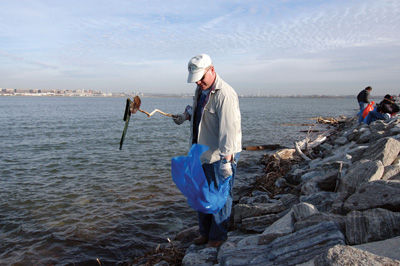 A man stands on the banks of a river and collects garbage into a plastic bag. In the background, others do the same thing.