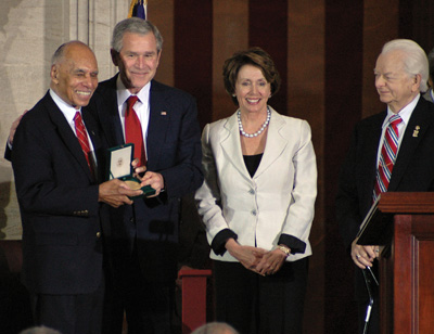 A man in a suit stands next to another man in a suit. Together they hold a medal in a small box. A woman and another man stands nearby.   