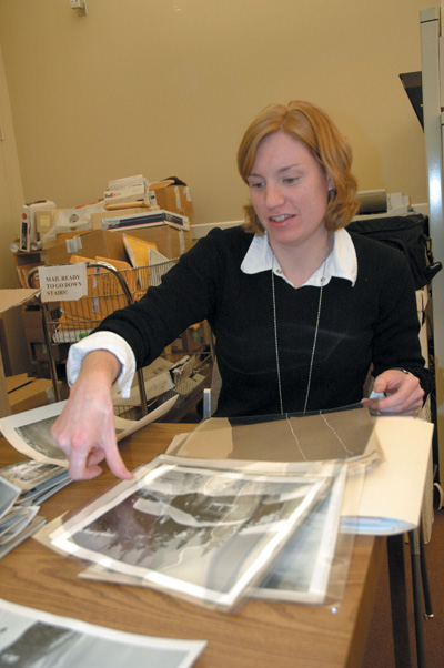 A woman sits at a desk that has folders and photographs on it. Behind her is a metal cart full of outgoing mail.