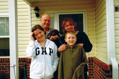 A man, a woman, a girl and a boy stand together in front of a home.