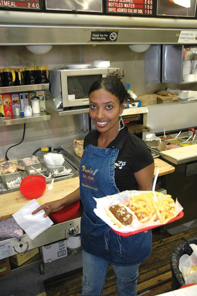 A woman holds a tray with a chili dog and French fries with cheese.  She is in a commercial kitchen. He apron says "Ben's Chili Bowl Washington D.C."  