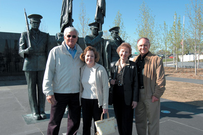 Four civilians stand in front of large bronze statues that depict men in military uniforms.  Trees are visible in the background.