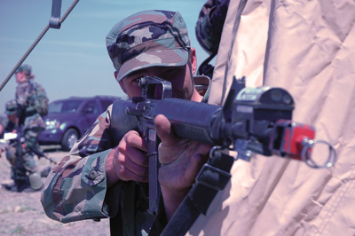 A man in a military uniform aims a rifle around a corner.