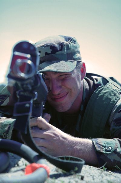 A man in a military uniform lays on the ground and aims a rifle.