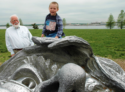 Outdoors, a small boy sits on what appears to be a part of a bronze sculpture. Grass and a river can be seen behind him. An elderly man stands nearby.