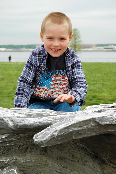 Outdoors, a small boy sits on what appears to be a part of a bronze sculpture. Grass and a river can be seen behind him.