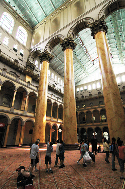 A huge indoor space has three large stone columns and glass ceilings and many arches lining the floors that surround the space.  People on the ground look upward.