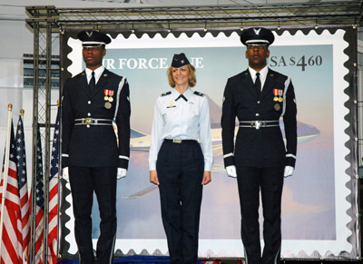 Two men in military uniforms flank a woman in a military uniform.  They stand in front of a large replica of a stamp that says "Air Force One USA $4.60."