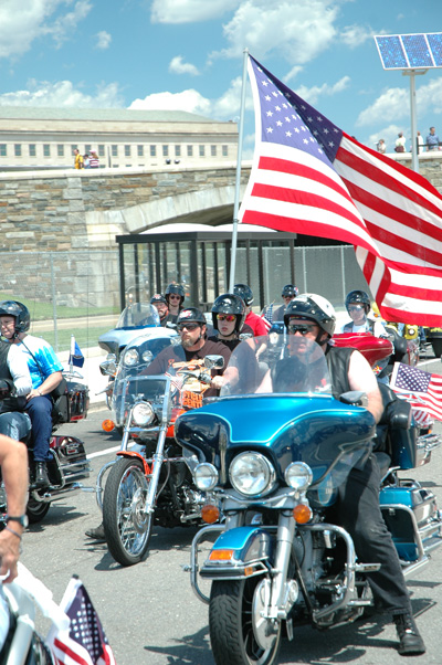 A procession of motorcycles.  Many have American flags. The Pentagon an be seen int he background.