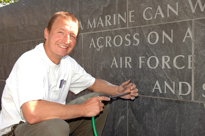 A man squats in front of a granite wall. The wall has words carved into it. he holds a green hose attached to a tool.  The words that are visible read "A Marine can ... across on a .. Air Force .. and."