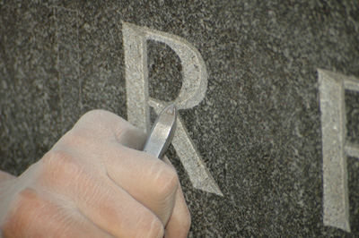 A hand holds a chisel and carves the letter "R" into granite.