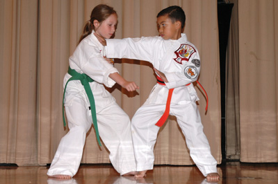Two children engage in martial arts. They each wear white outfits. The girl on the left has a green belt, the boy on the right has a red belt.