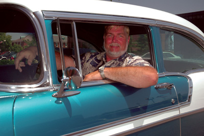 A man sits in an antique car. His elbow is hanging out the driver's side window.