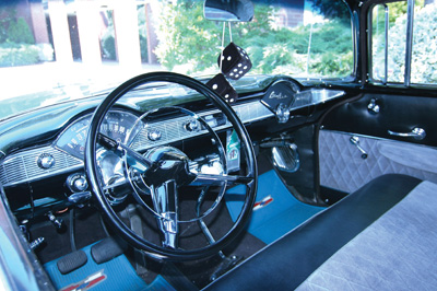 The inside of an antique car has blue carpet and a lot of chrome. Fuzzy dice hand from the rear-view mirror.  