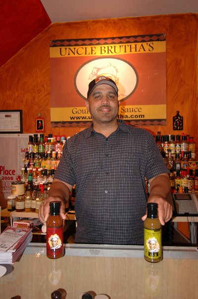 A man stands behind a counter.  Two bottles of hot sauce are on top of the counter. Behind him is a sign that says "Uncle Brutha's," and also shelving with dozens of bottles on it.