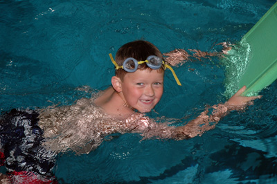 A small boy swims in a pool. He holds a kick board and wears goggles on his head.