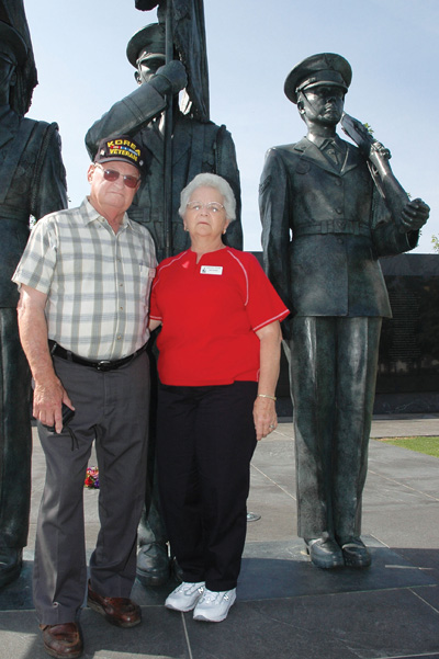 A man wearing a hat that says "Korea Veteran" and a woman stand together in front of three large bronze statues of uniformed men.