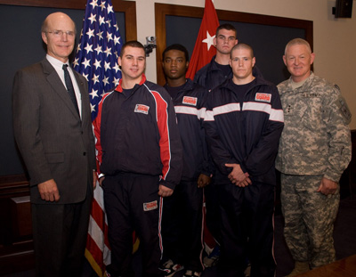 A man in a suit, four young men in track suits, and a man in a military uniform stand together in a room.  Behind them are the American flag and a general's flag with three stars on it.