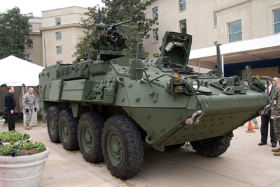 A military combat vehicle sits on a cement patio in a courtyard.  People stand nearby.  The vehicles front hatch is open.