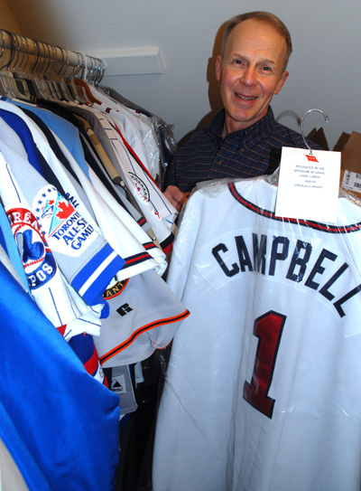 A man holds a sports jersey that says "Campbell 1" on the back.  Hanging up on the rack to his right are many other similar jerseys.