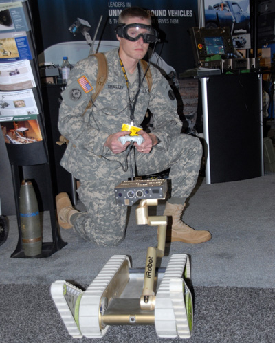 A soldier is down on one knee in a carpeted room. He wears eye protection and hold a remote control. in front of him is a tracked robot that says "iRobot" on it.  behind him are various displays and racks with reading material.